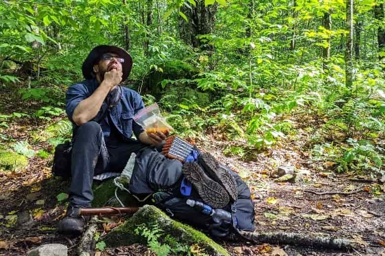 backpacker sitting on the ground with a bag of snacks eating. 