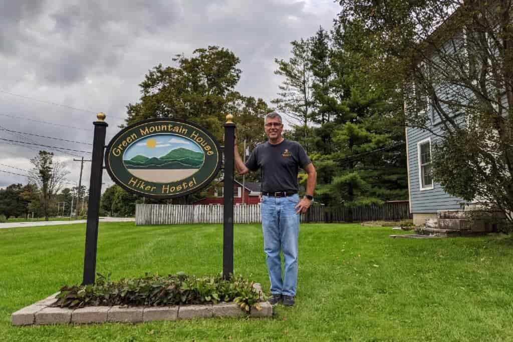 hiker hostel owner posing next to the hostel's sign on a grassy lawn. 