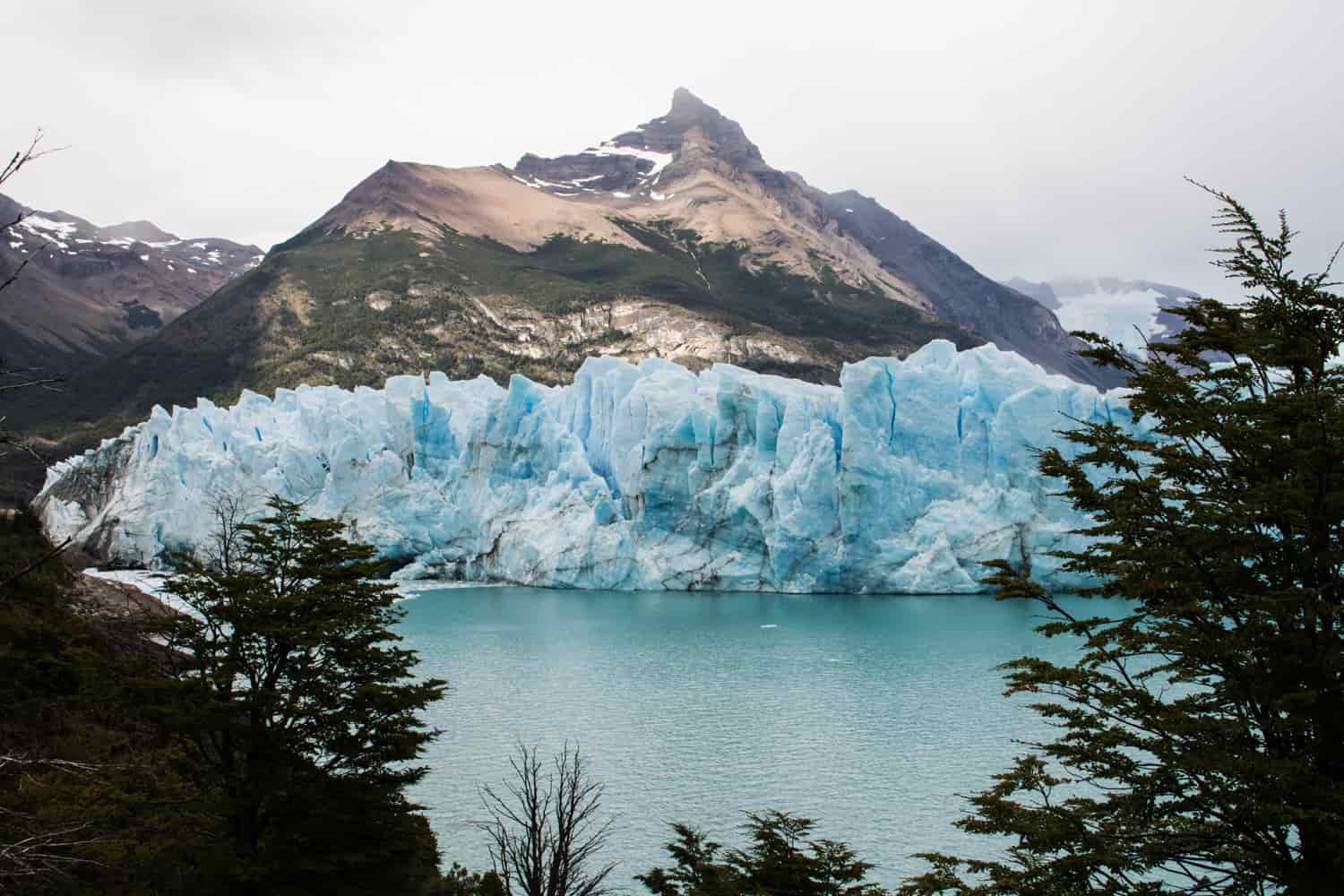 The blue-ish ice wall of a glacier sits on the edge of an alpine lake