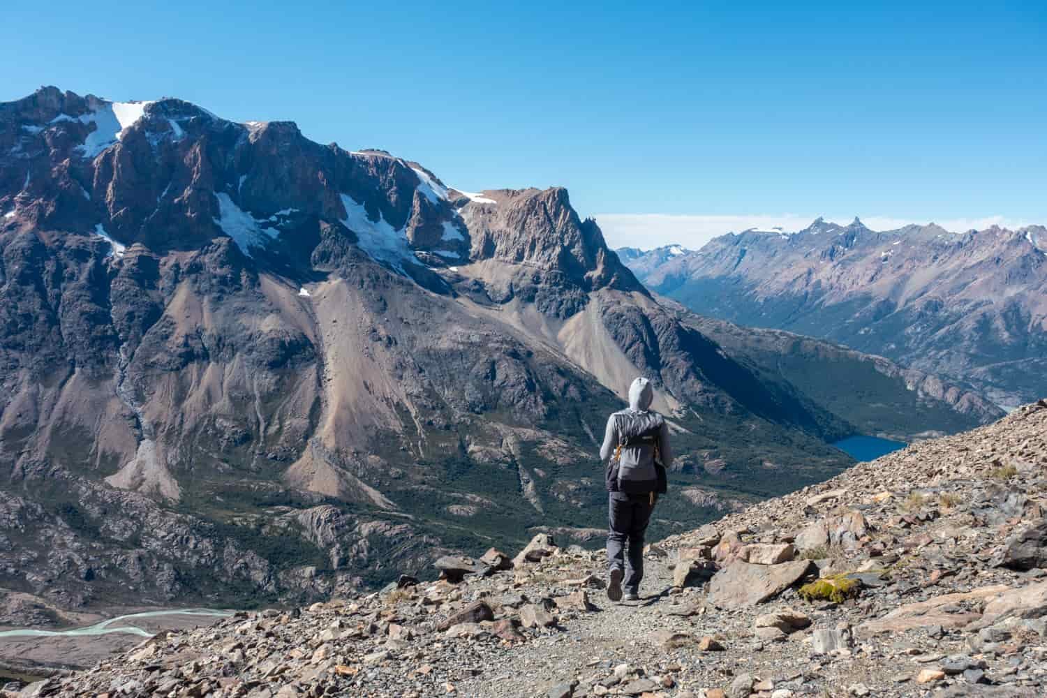 A female hiker walks down a trail with big views of the mountains above her and valleys beneath her