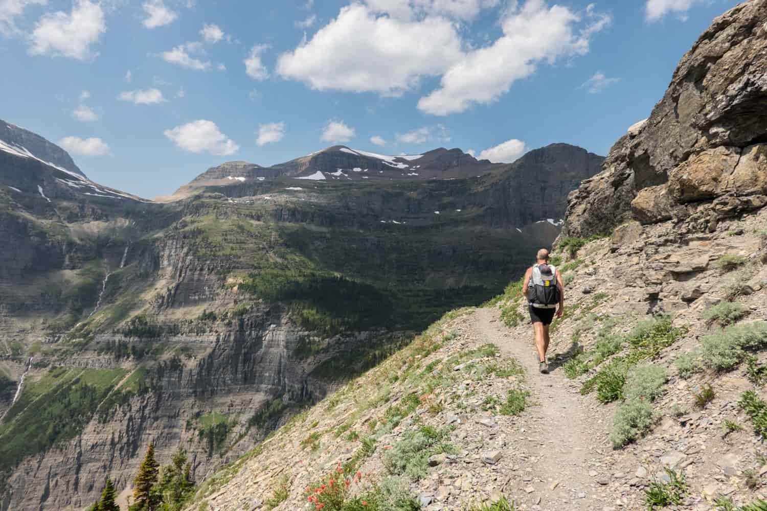 waterfall and high alpine creek cascading in the distance while a backpacker hike a rocky trail in glacier national park