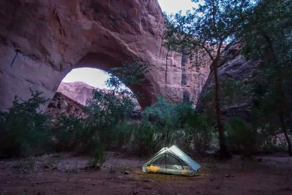 a tent with a light in it below a large rock arch in the desert