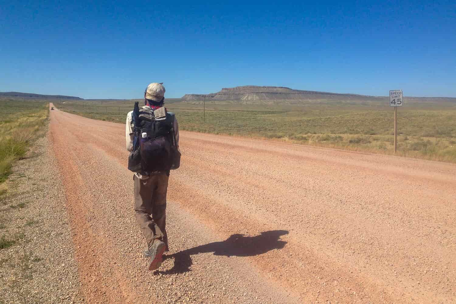 CDT thru-hiker walking along a straight dirt road in the sun with a cloudless blue sky.