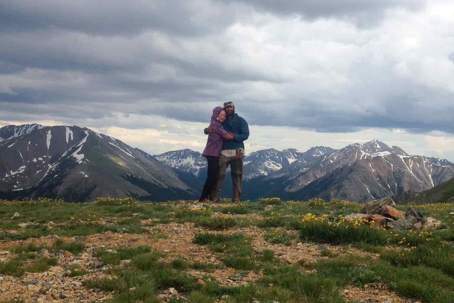 Two thru-hikers stop for a hug and picture on top of a beautiful mountain view on a cloudy day.