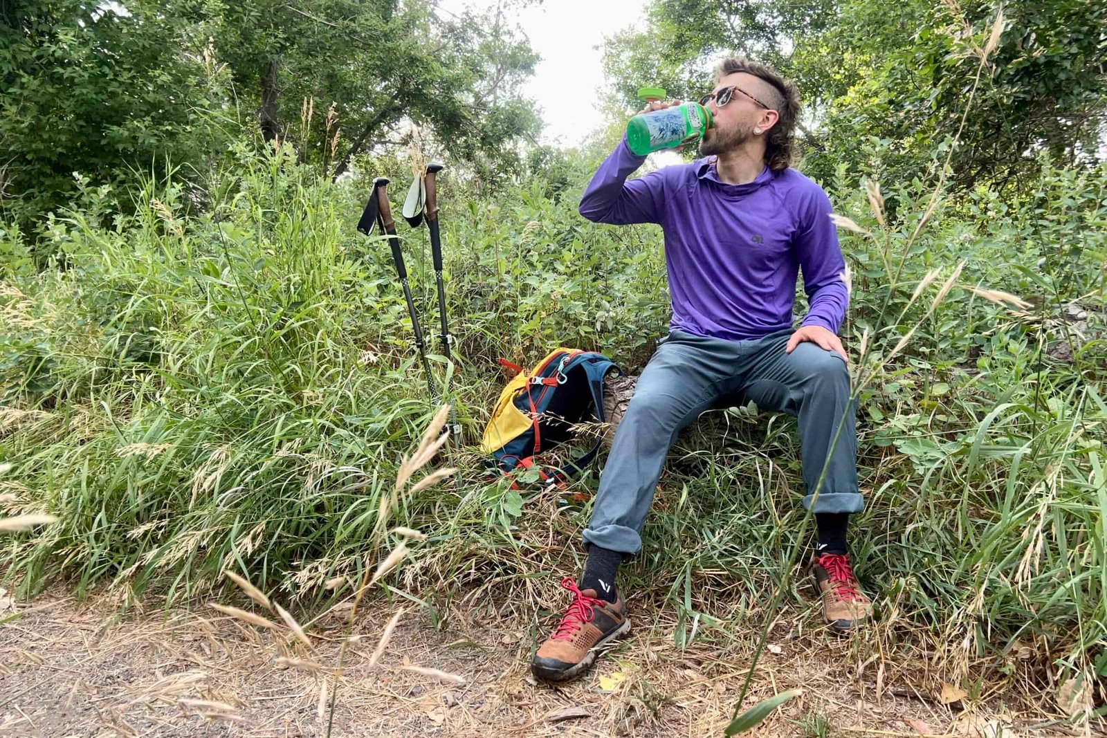A man sits on a log drinking water on a hike with his pack and poles next to him.