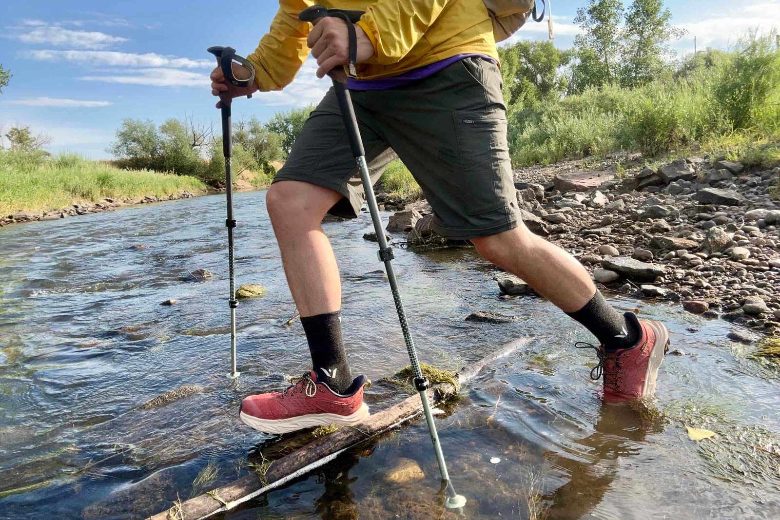 A male hiker walks across a river in a wilderness area using poles.
