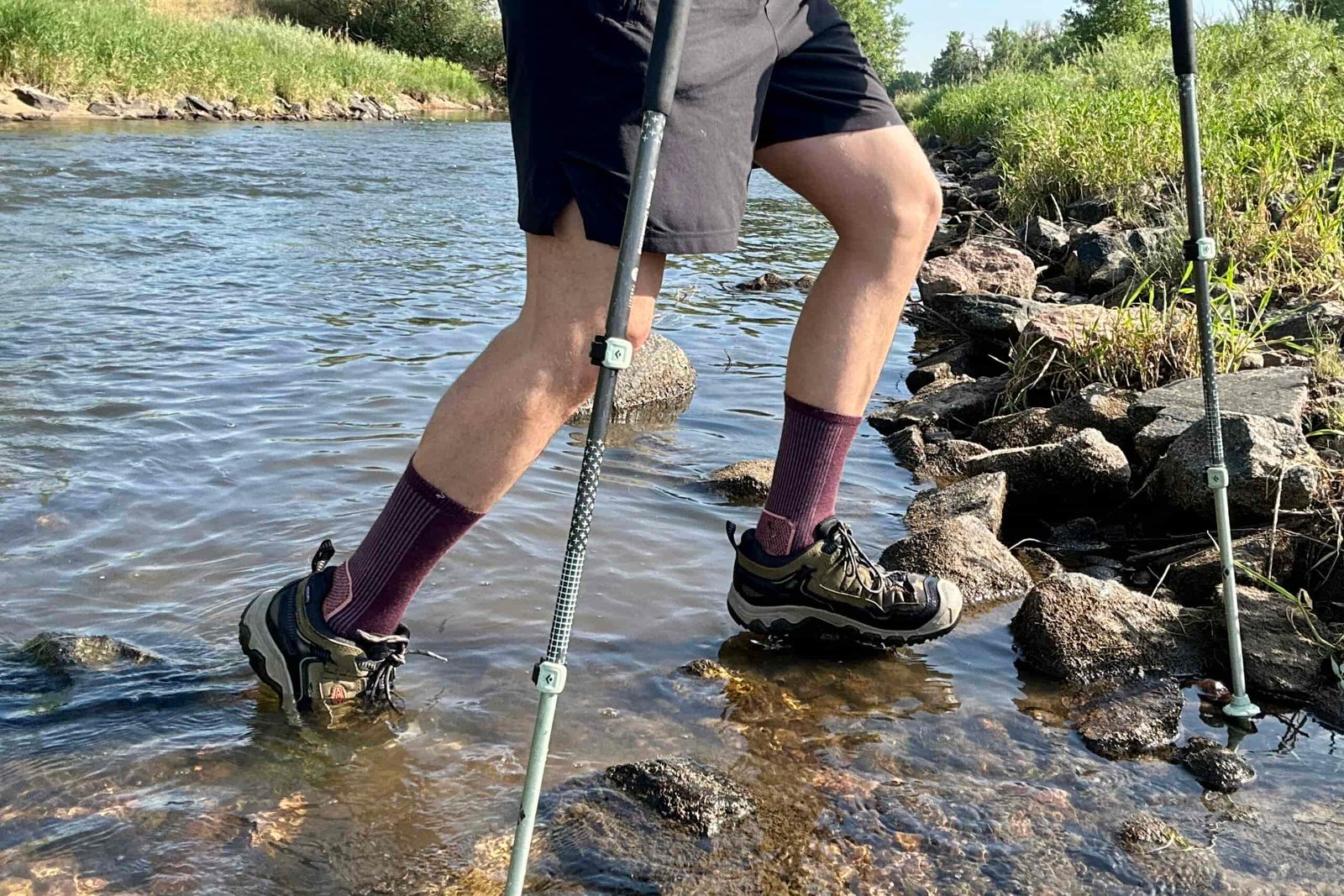 A male hiker crosses a river. The picture is from the waist down.