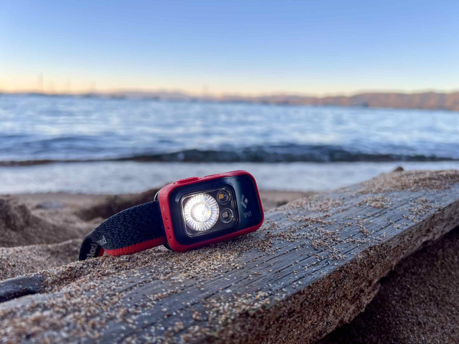 The image shows the Black Diamond Spot 400 sitting on a beach with it's floodlight illuminated. There is water in the background.