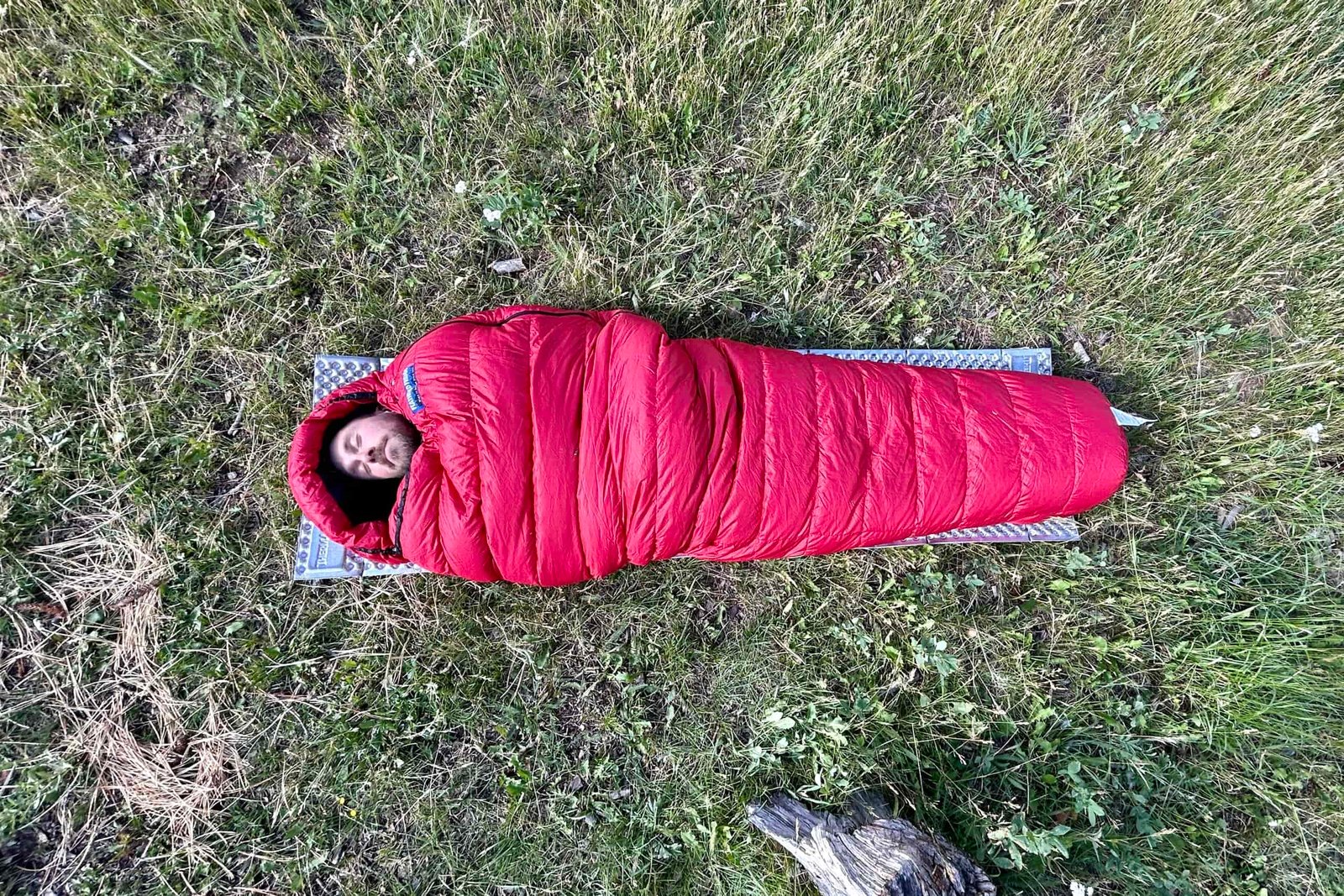An mage from above of a man resting in a sleeping bag on a foam pad with the hood up. He is cowboy camping on the grass in the wilderness.