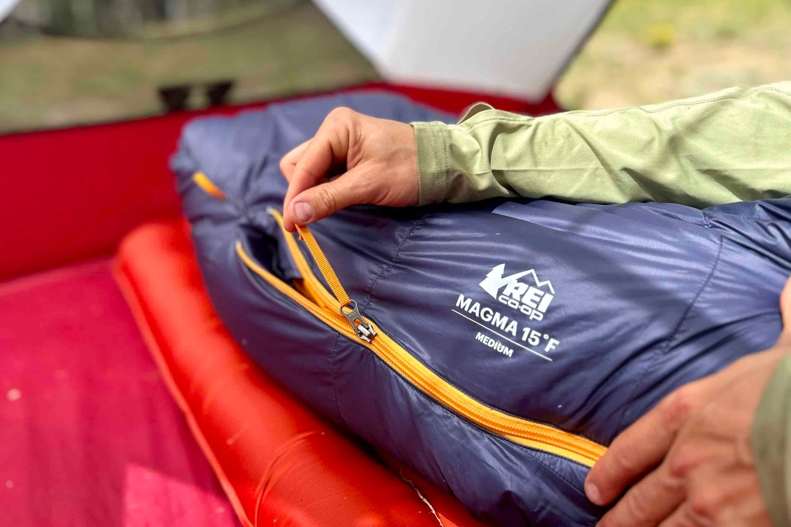 A person zips up a sleeping bag inside a tent. Only their hands and the footbox of the sleeping bag are visible.