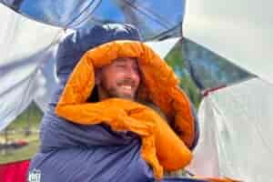 A man sits up in a sleeping bag with the hood over his head. He is smiling and looking off camera.