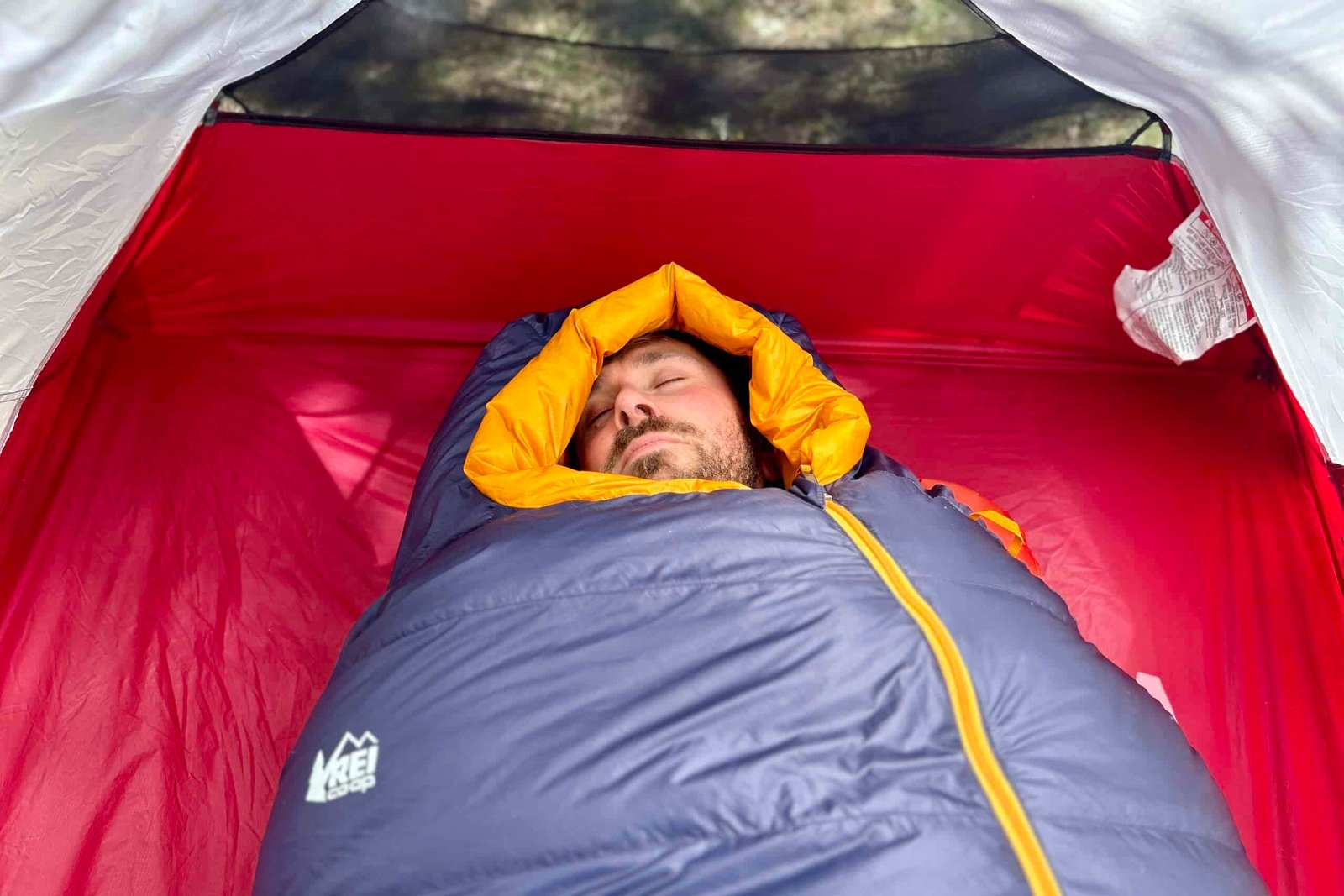 A man in a sleeping bag rests with the mummy hood cinched around his face in a tent.