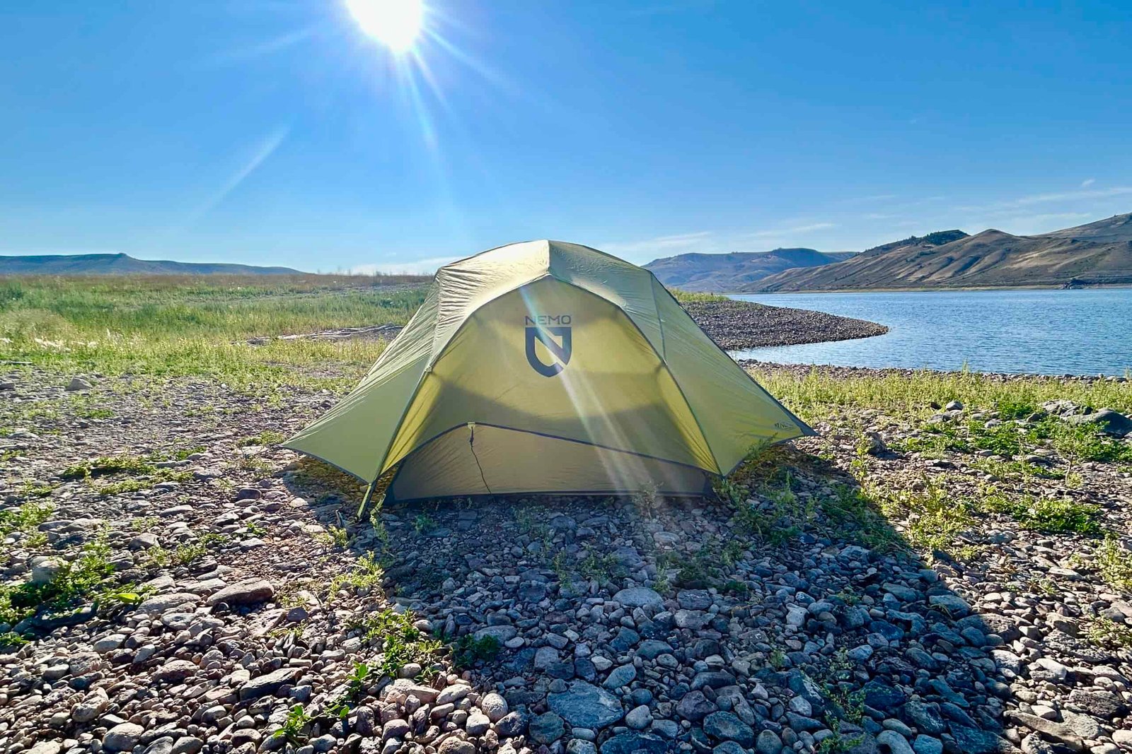 A tent stands alone in the center of a rocky beach with a lake in the background.