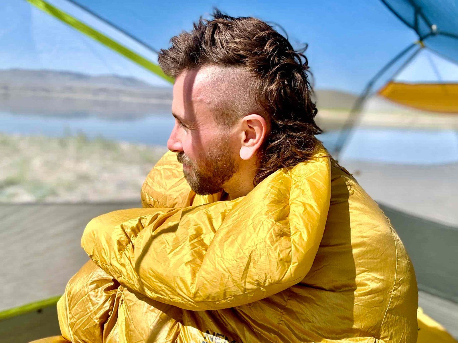 A close up of a man from the waist up, sitting up in a tent and looking off in the distance, with a sleeping bag pulled around his shoulders. There is a body of water and mountains blurred in the background.