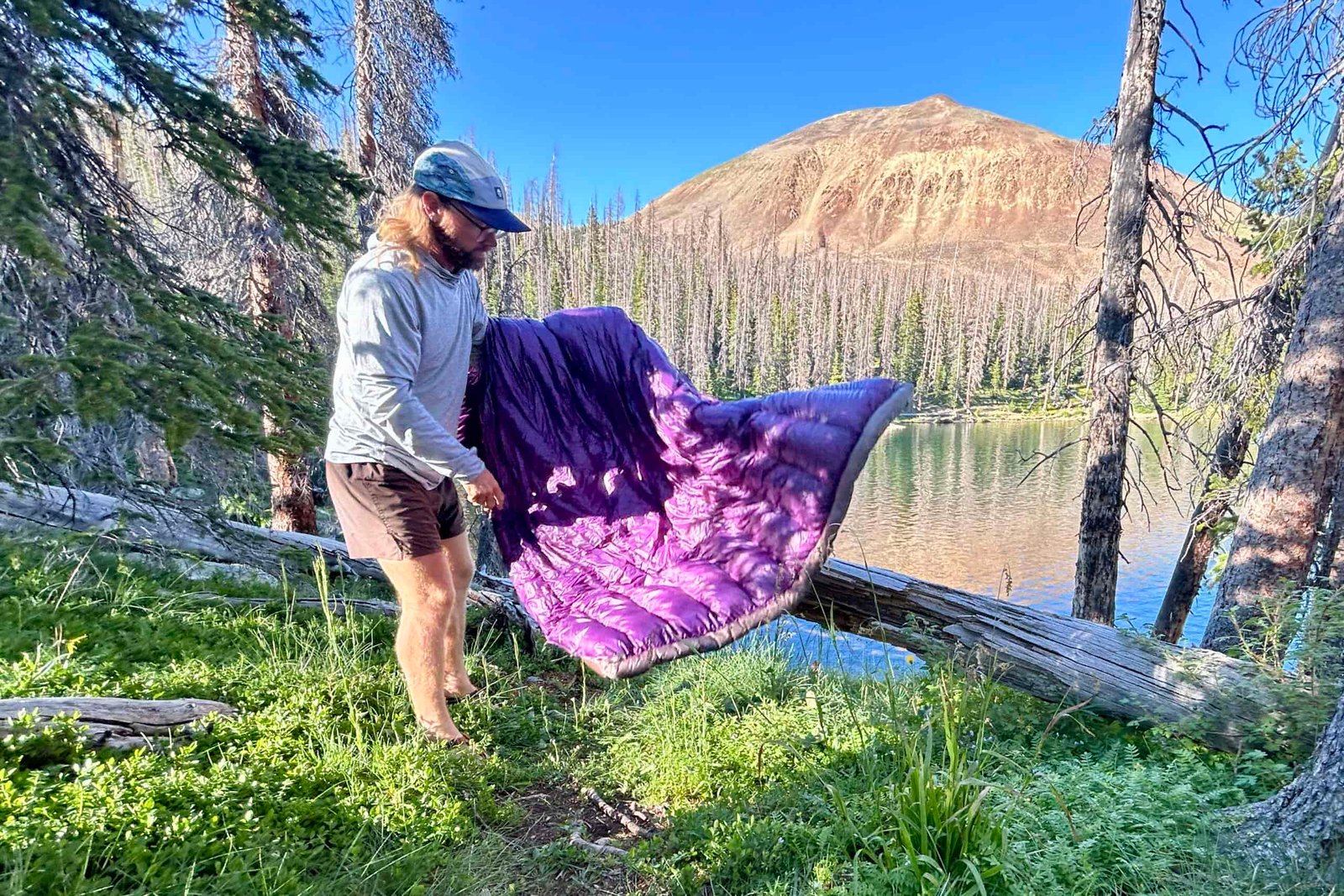 A man stands on the banks of a large alpine lake shaking out a purple quilt.