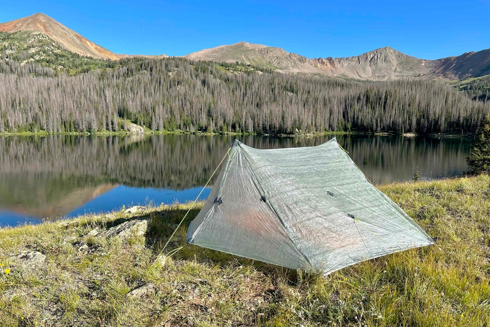 the zpacks duplex classic tent pitched near a glassy lake on a bright cloudless day. redish brown mountains enclose the lake