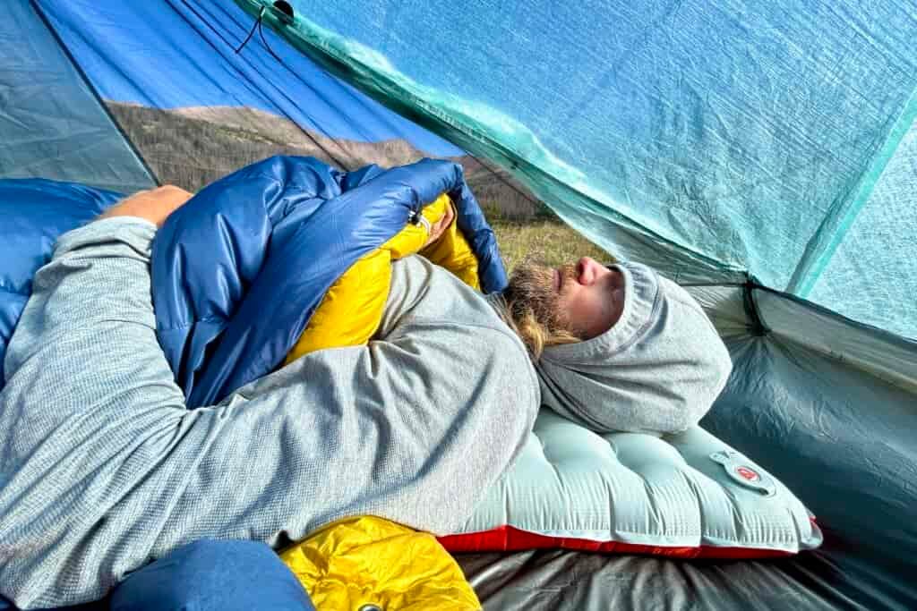 A profile image from the waist-up of a man with his hood up, eyes closed, and arms out and sleeping quilt pulled up underneath his arms.