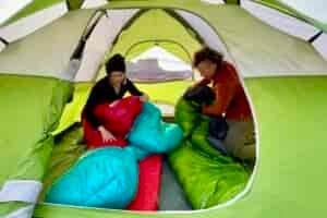 Two people pack up their sleeping bags in a green tent with a large desert butte visible in background.
