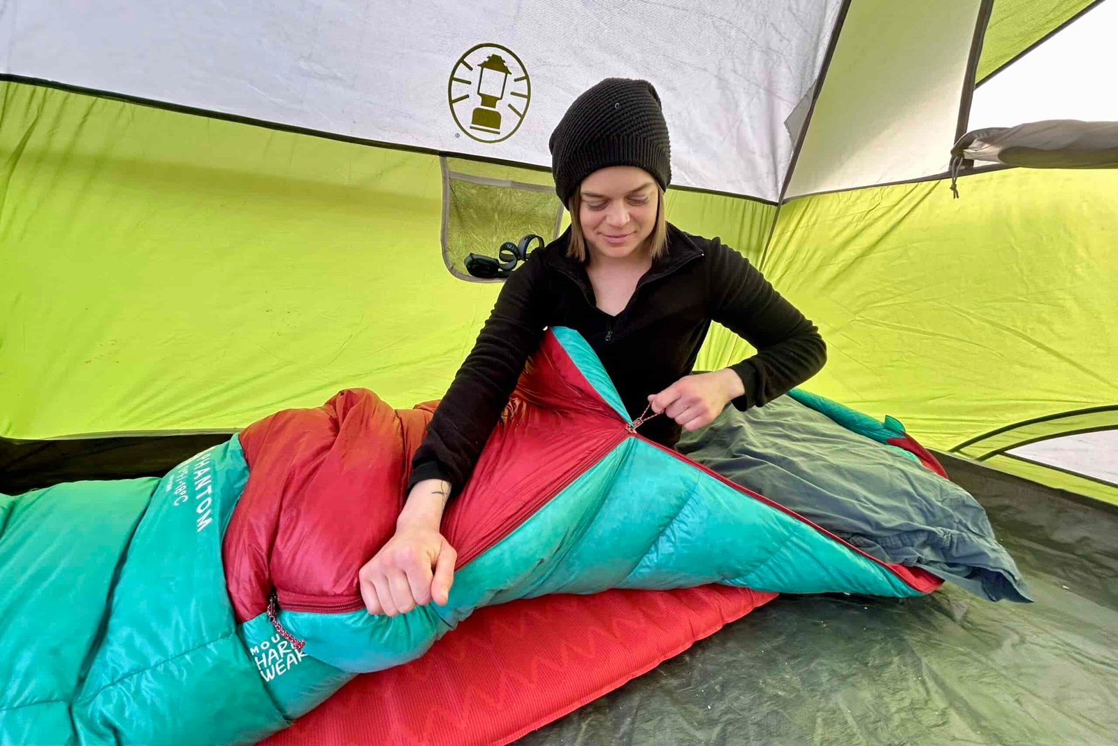 A woman wearing a beanie zips up a sleeping bag while sitting inside it.