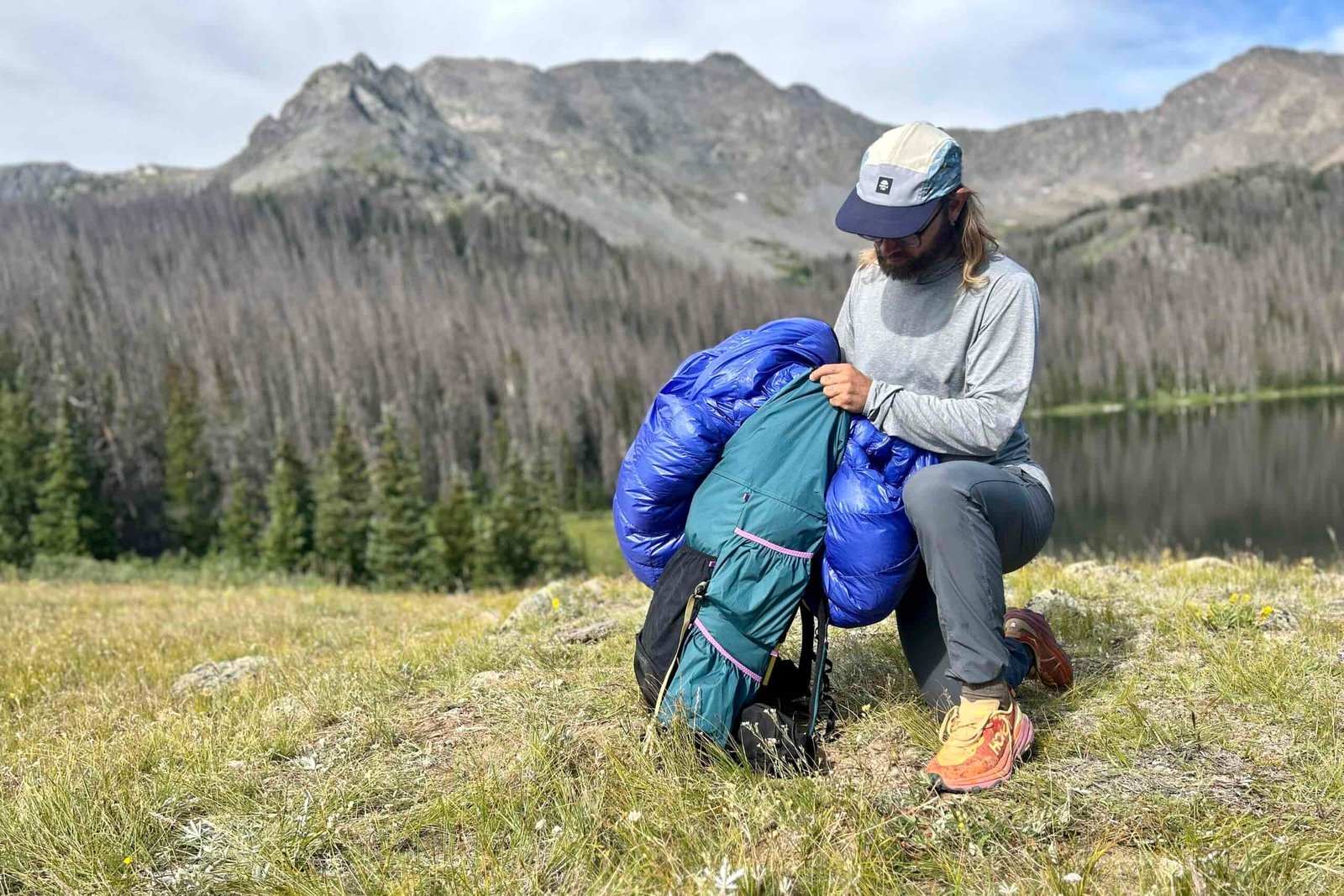 A man stuffs a sleeping bag into a backpack next to an alpine lake with mountains in the background.