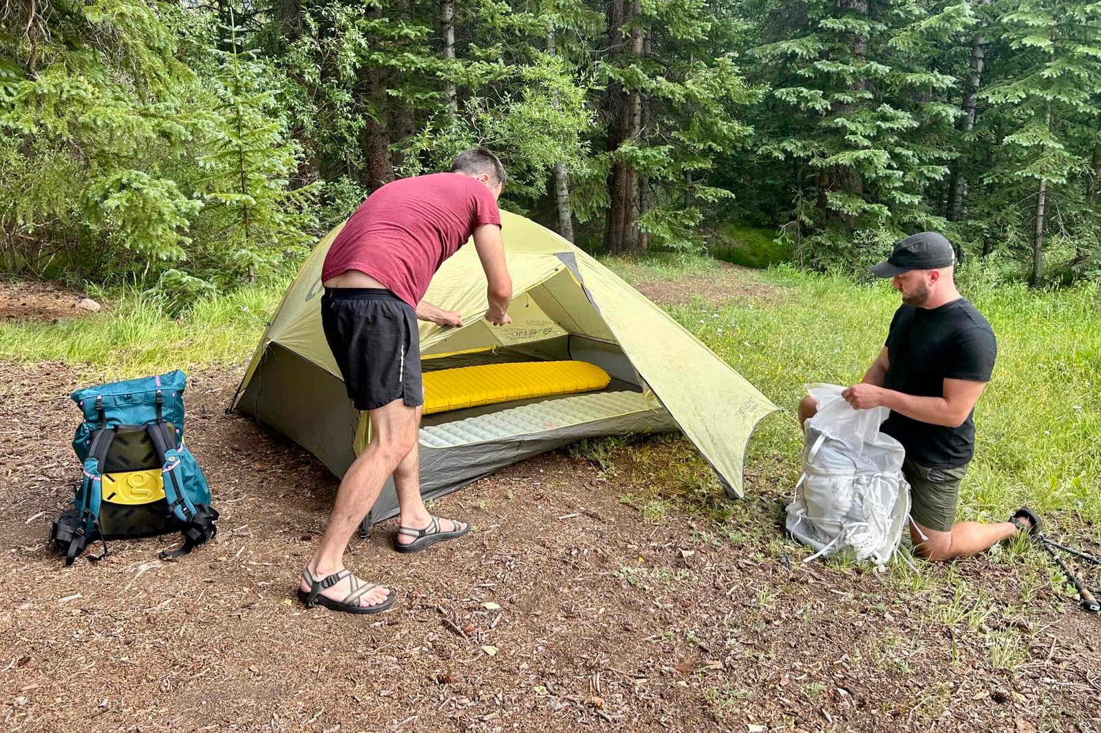 2 men in the woods unpack their backpacks and set up a tent with the rainfly.
