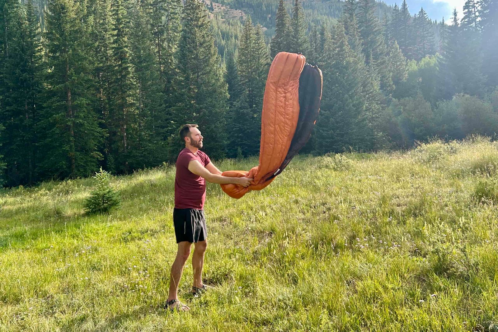 A man shakes out a sleeping bag in a grassy field on a hot summer day.