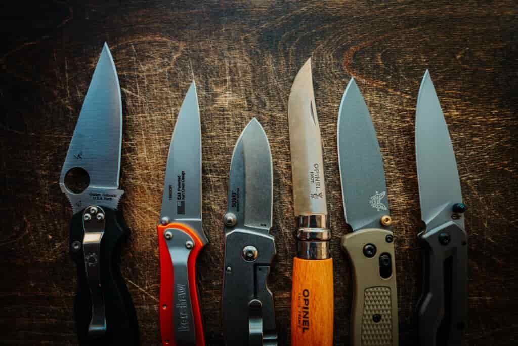 Six pocket knives are laid out on a wooden background with blades open to show blade length and shape.
