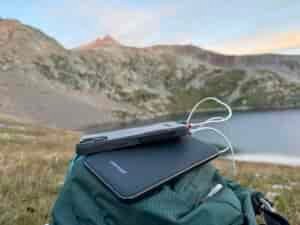 Close up of the Charmast power bank charging a phone that is sitting on a backpack with a lake and mountain in the background.