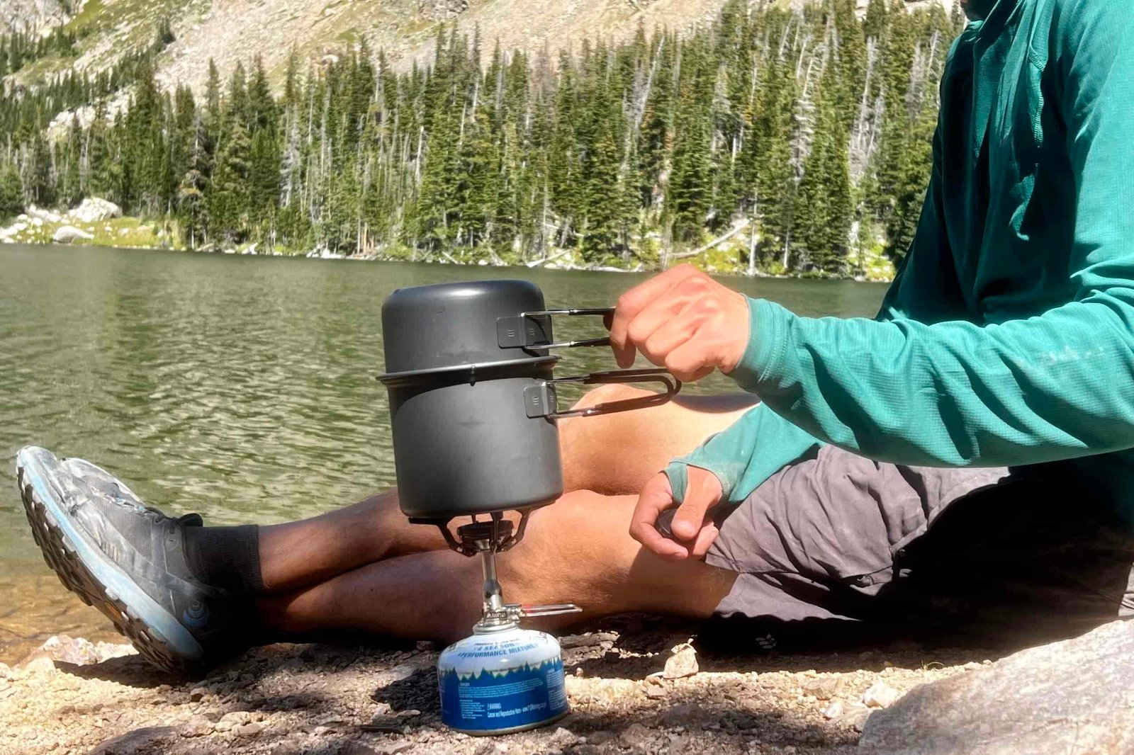 A hiker cooks with the SOTO Amicus Cookset Combo. The stove and fuel canister sits next to him, with the pot on the flame. He is placing the lid of the cookset combo on the main pot. There is a lake and trees in the background.