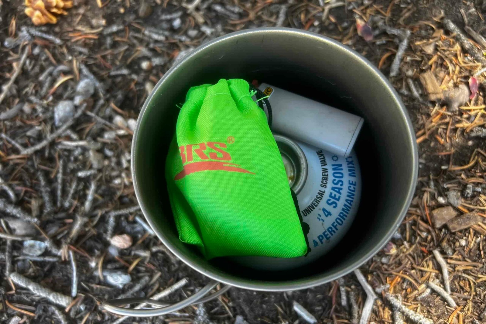 The BRS 3000T stove packed in a 550mL pot with stuff sack, an 8oz fuel canister and mini lighter. The whole kit sits on a pine needle forest floor.