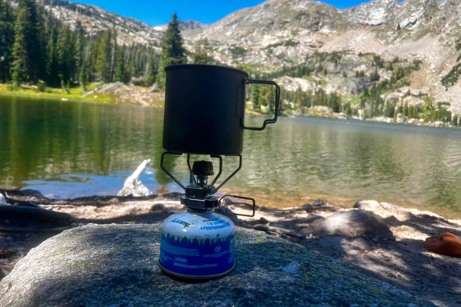 The Snow Peak Gigapower 2.0 sits on a rock, installed on a fuel canister, with a pot on top. The background is mountains and a lake.