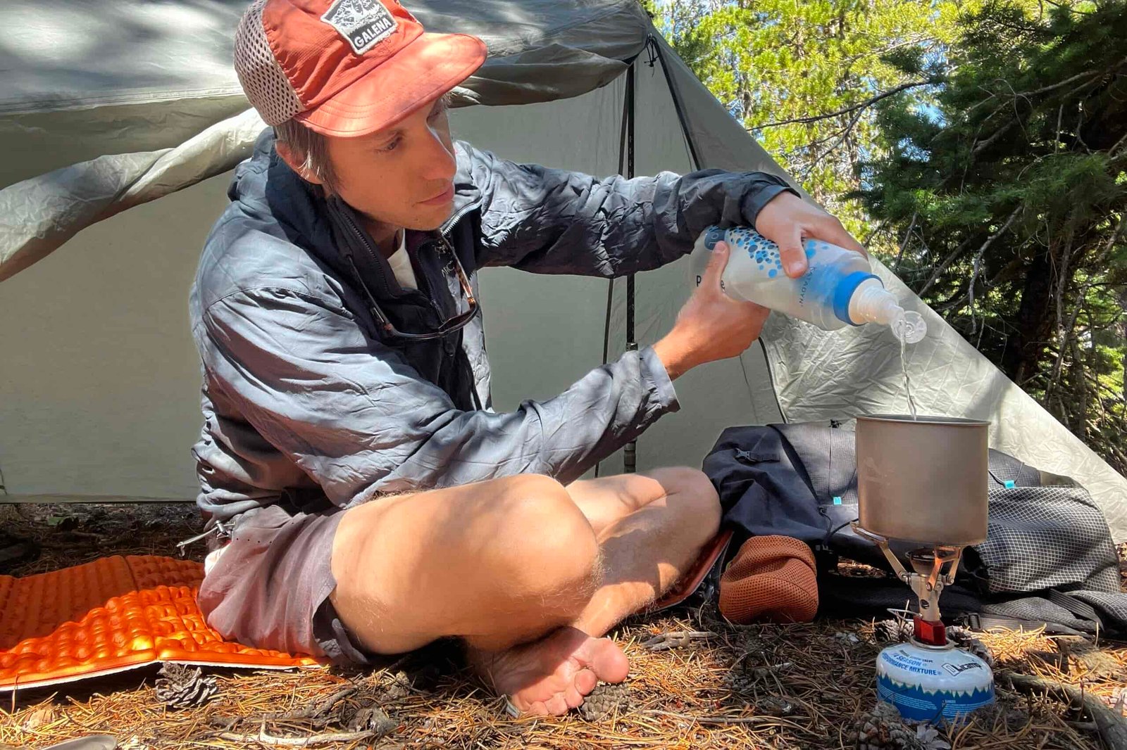 A hiker filters water into a pot that is sitting on the MSR PocketRocket 2. Tent, backpack, and trees are in the background.