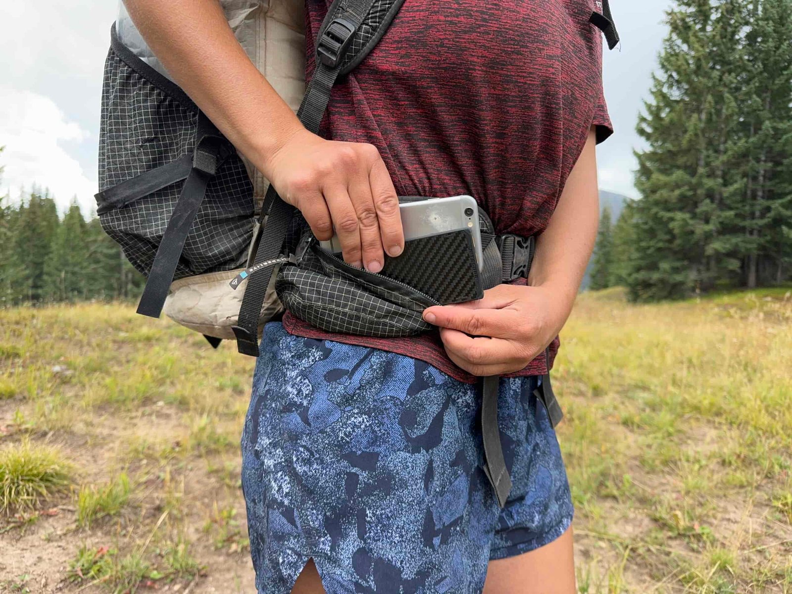 Close up of a backpack side pocket with one hand placing the Nitecore and a cell phone into the pocket and the other holding the zipper. There are trees in the background.