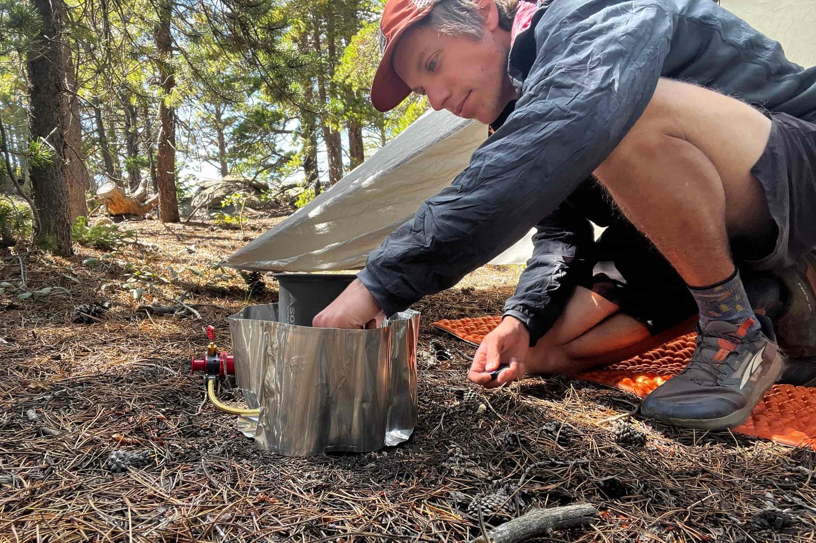 The Whisperlite sits on the ground with a pot on top and its windshield wrapped around. A hiker reaches down to light the priming cup of the stove. A tent and trees are in the background.
