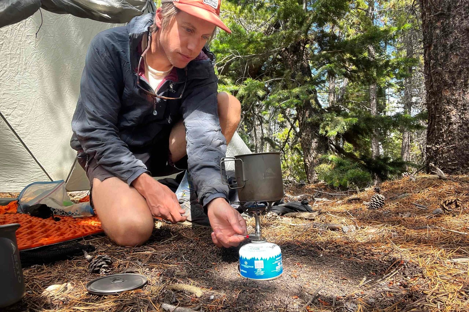 A hiker cooks with the SOTO Amicus Cookset Combo in a wooded, backcountry setting. He is adjusting the flame level on the stove.