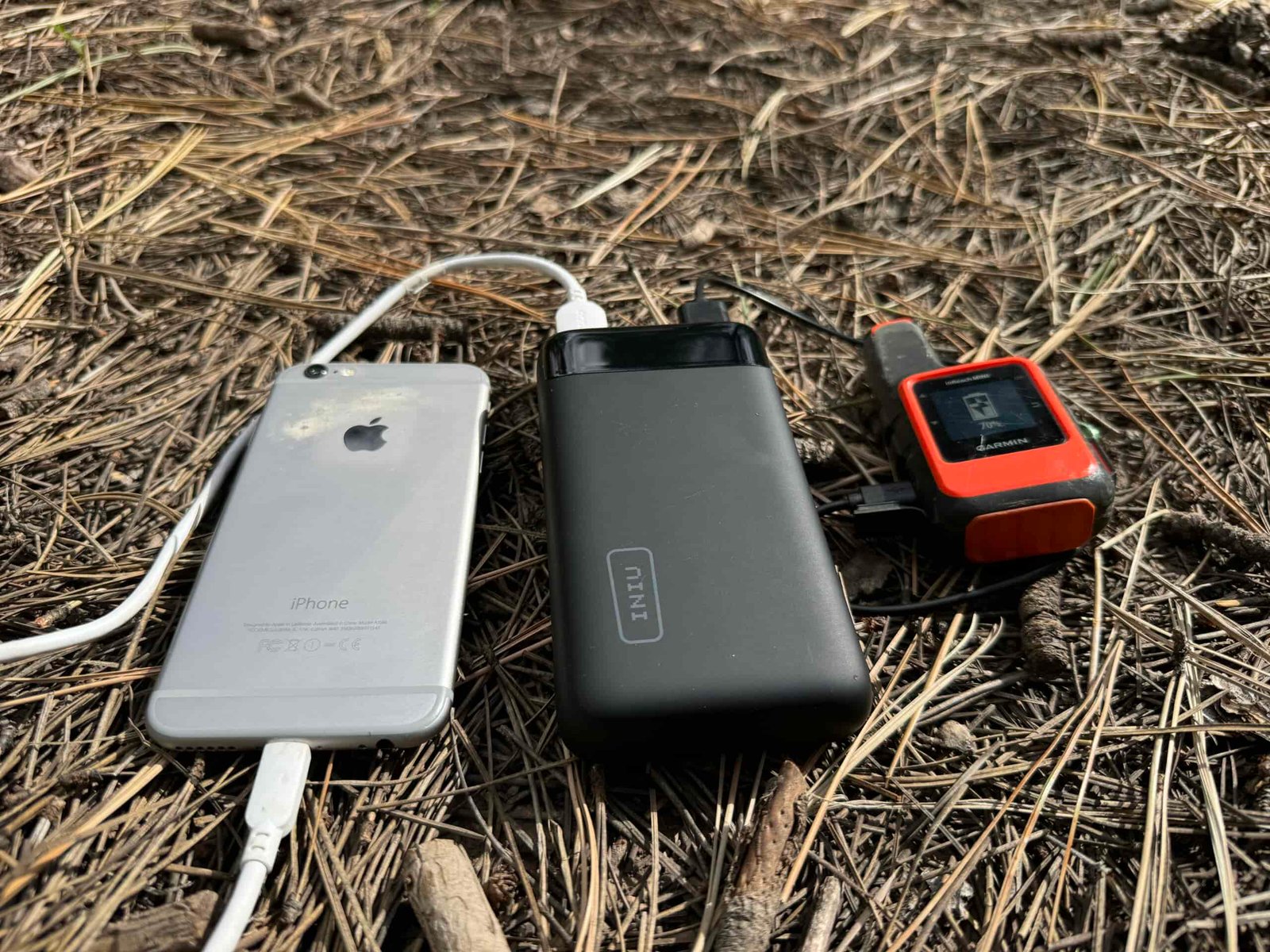 Close up of INIU 20000 PD power bank charging a phone and the Garmin Inreach while sitting on some pine needles.