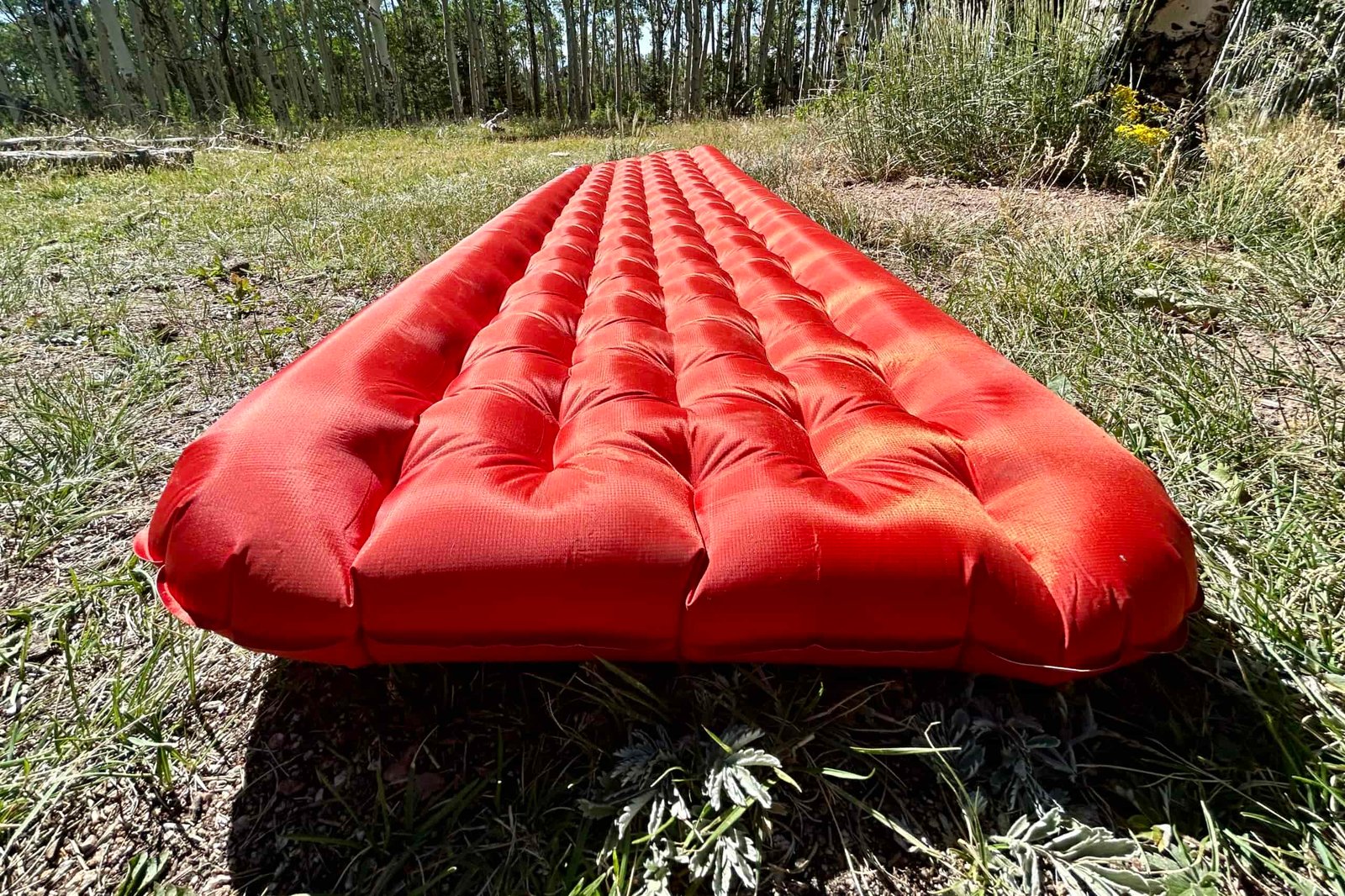 A red inflatable sleeping pad is laying on grass, showcasing its raised side rails and thick cushioning for support.