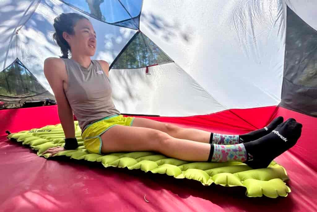 A woman relaxes inside a tent, sitting on a green inflatable sleeping pad. Trees are visible through the tent.