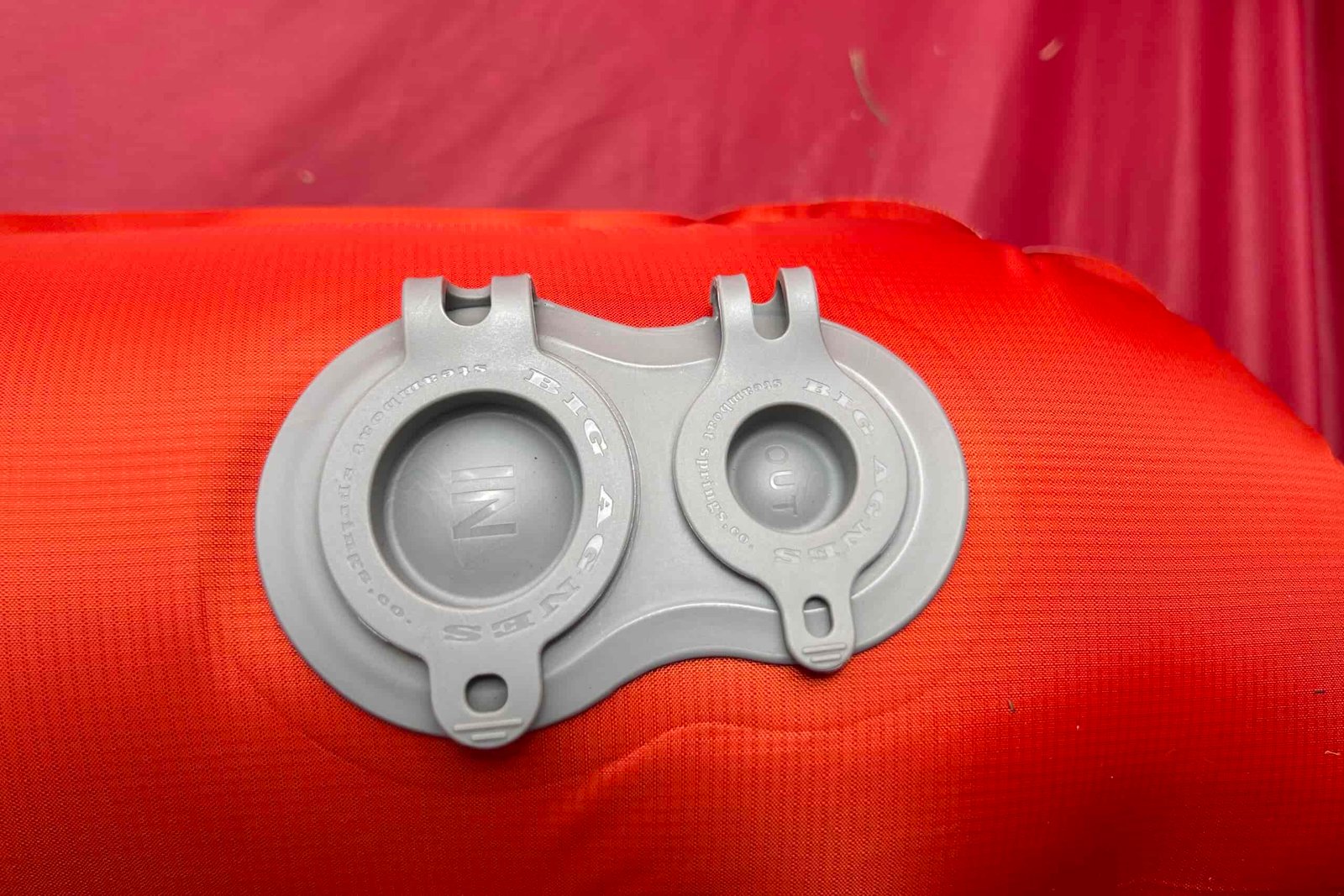 A close-up of a red inflatable sleeping pad with two gray valve caps, one labeled "OUT" and the other "IN", resting on grass.