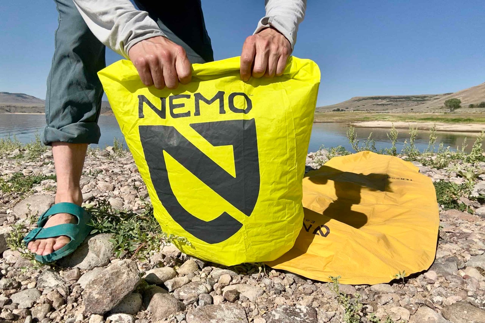 A person uses a bright green pump sack to fill an orange sleeping pad on a lakeside.