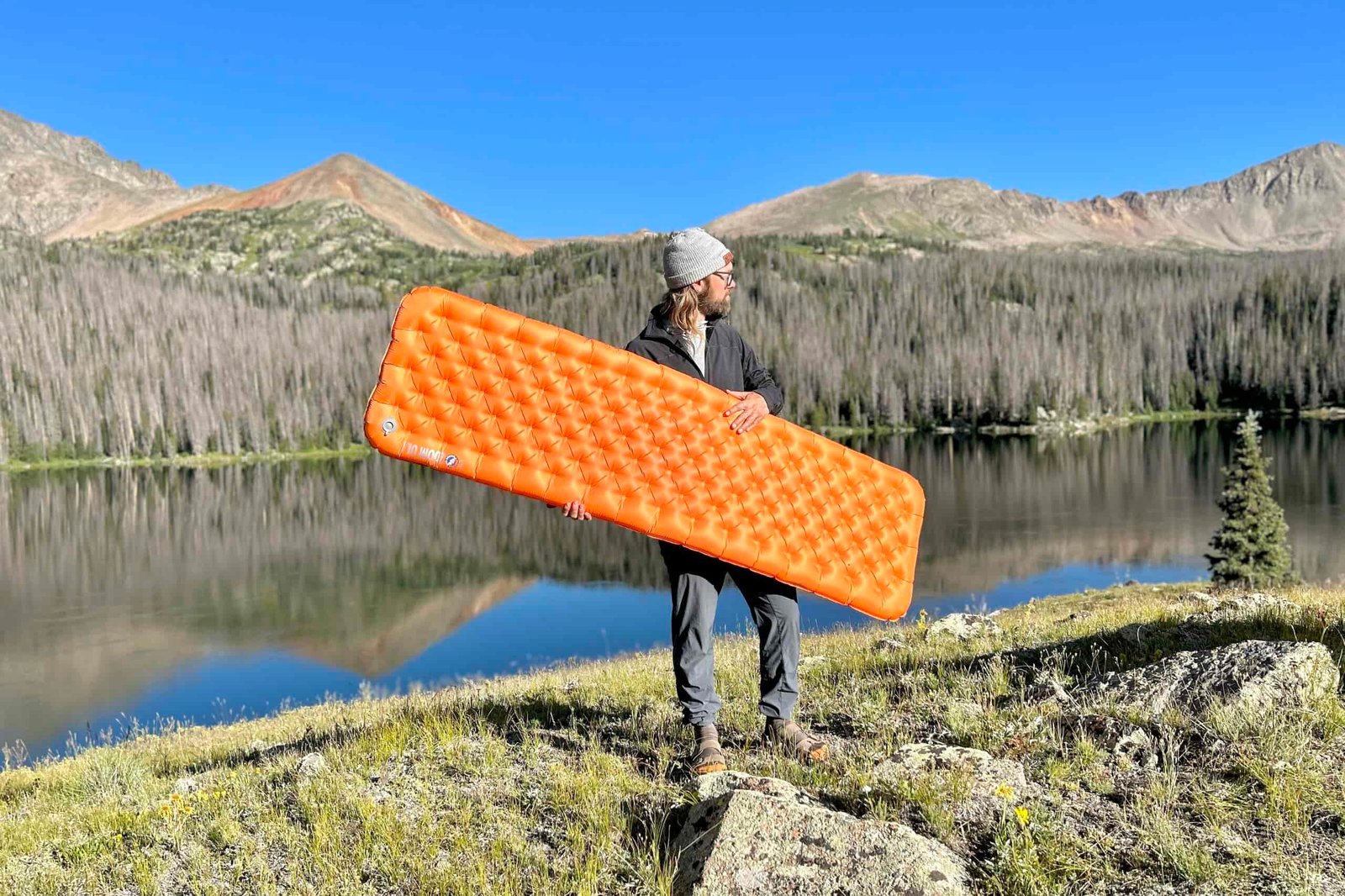A man stands in front of a lake and mountains holding an orange inflatable mattress.