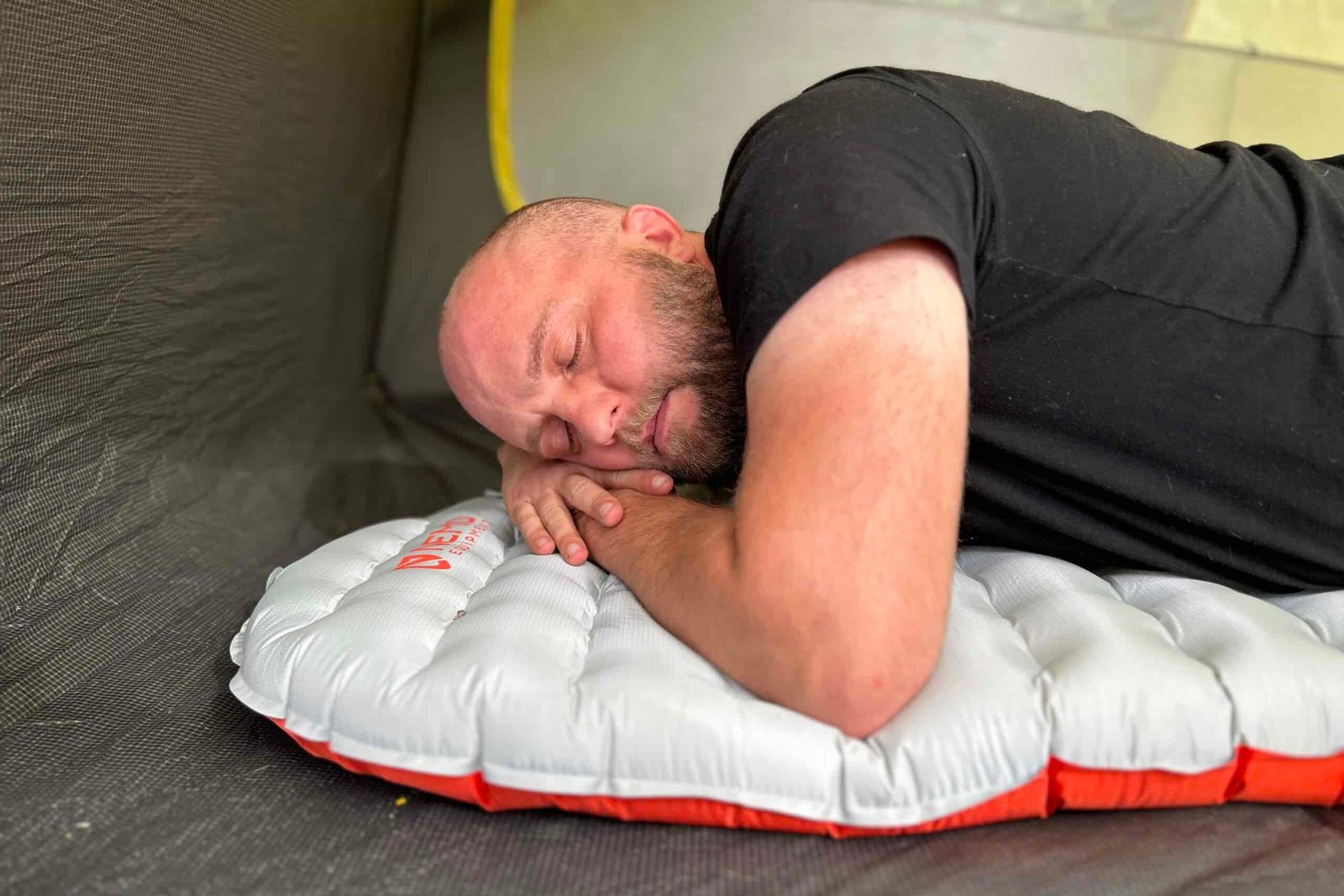 Inside a tent, a bald man lays on his stomach with hands under his head on an inflatable pad.