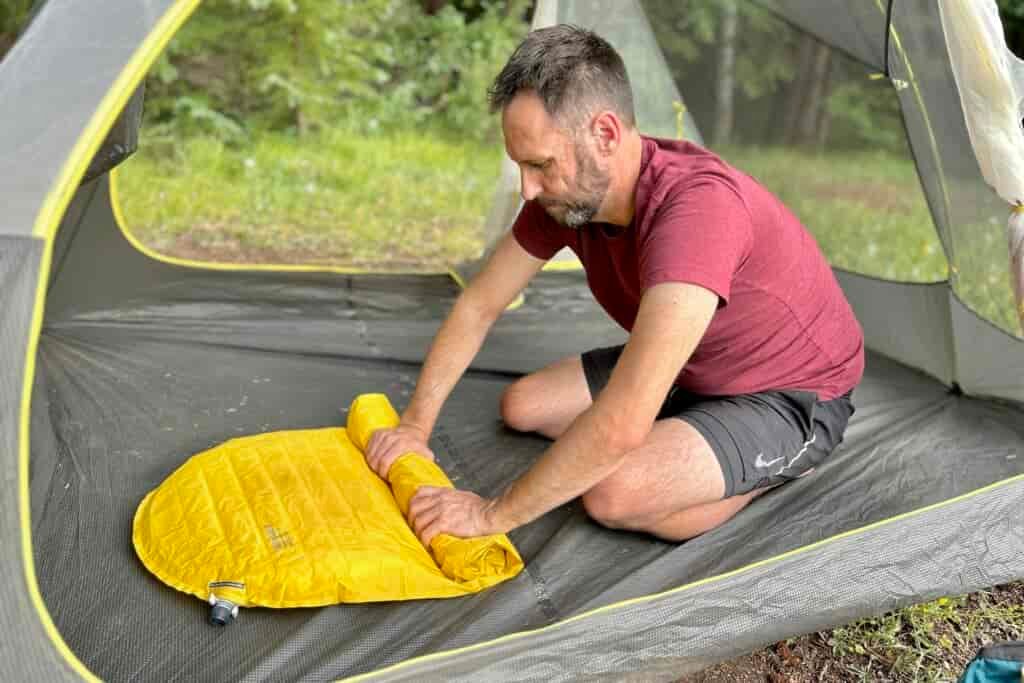 A man rolls up an inflatable sleeping pad in a tent.