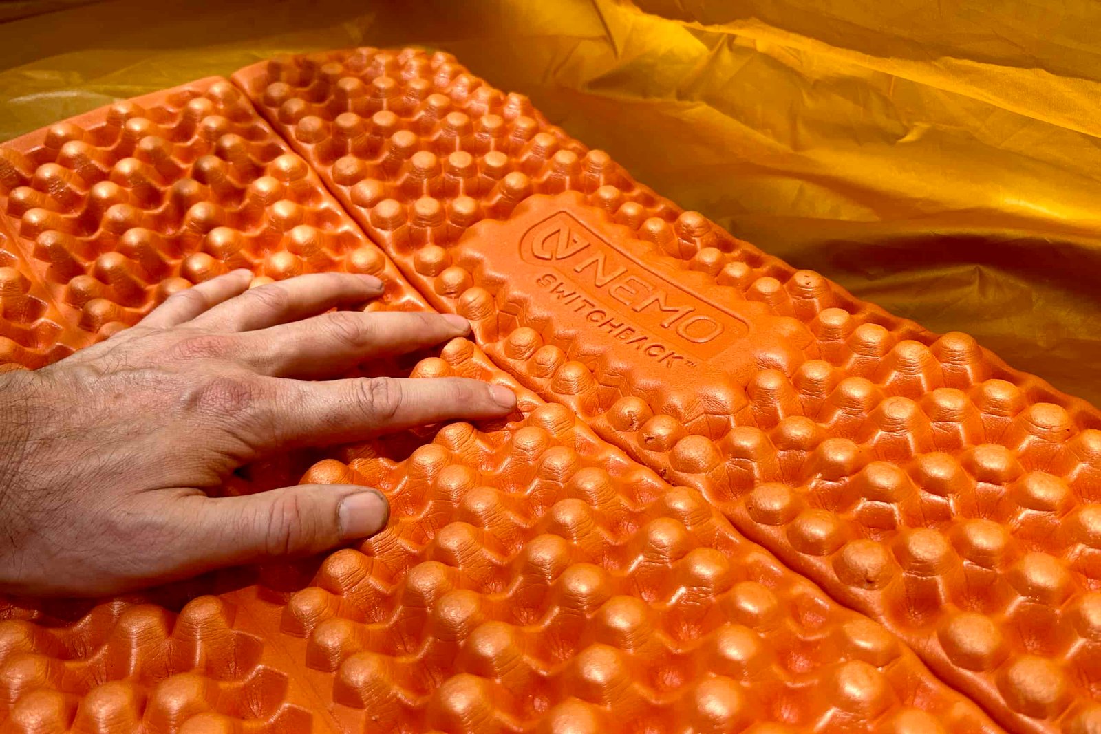 A person rests their hand on an orange foam sleeping pad in a tent.