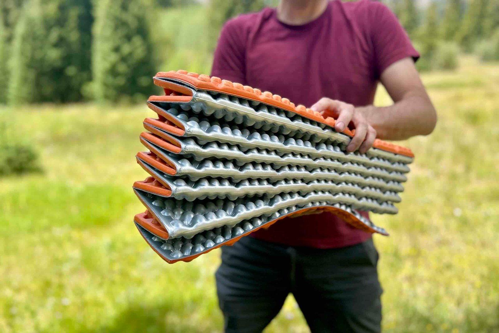 A man stands holding a vibrant orange and grey foam sleeping pad, showcasing the pattern and accordion-style packed size.