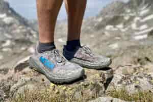 Hey side facing view of the HOKA Speedgoat 6 trail running shoes with a hiker, wearing them at high altitude with snowy peaks in the background.