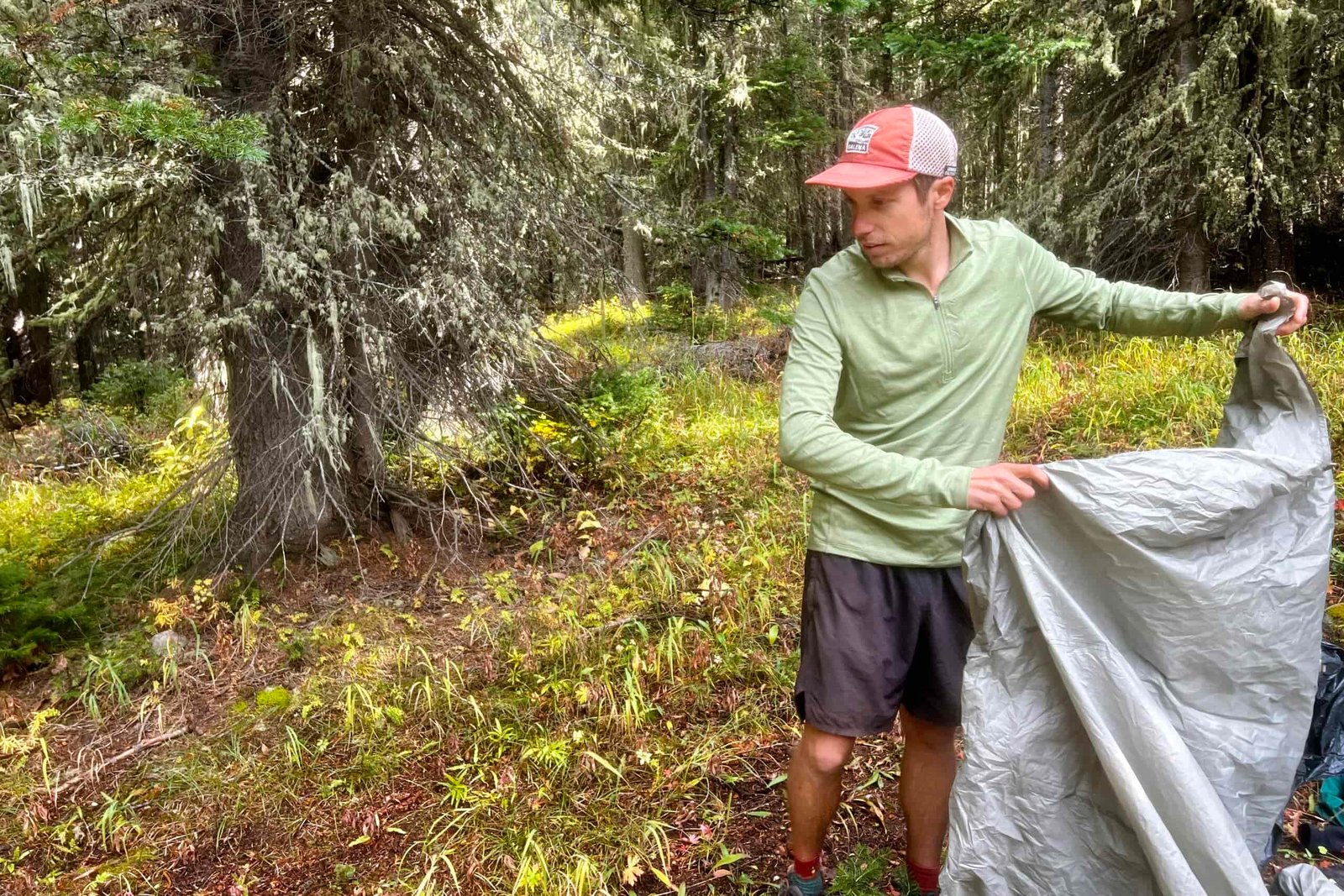 A hiker prepares to pitch his tent while wearing the REI Midweight Half Zip base layer. Trees are in the background.