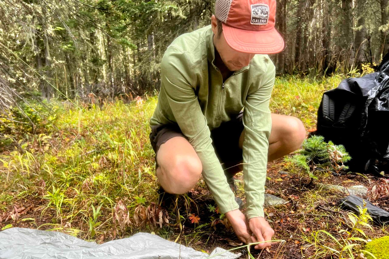 A hiker crouches to install a tent stake while wearing the REI Midweight Half Zip base layer. Trees and backpacking gear are in the background.