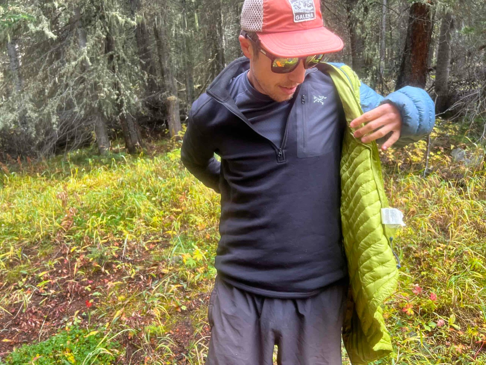 A hiker puts on a puffy jacket over top of the Arc'teryx Rho Heavyweight Zip Neck base layer. Trees are in the background.
