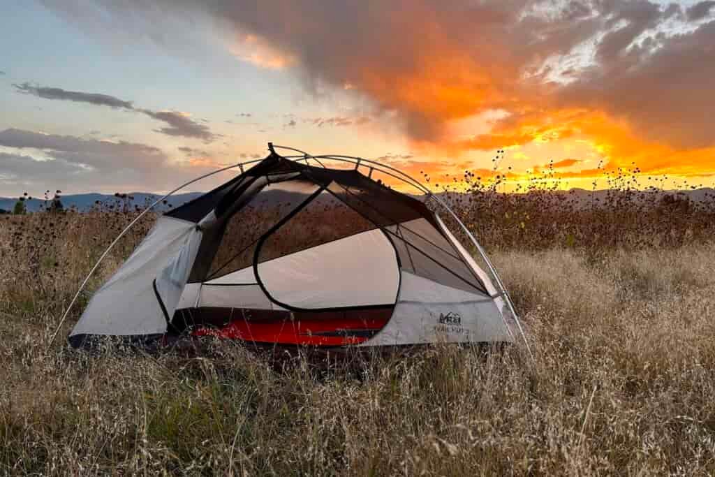 A tent with no fly on sits in a grassy field with a sunset in the background.
